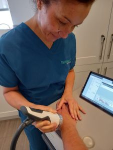 Medical professional in blue scrubs applies a handheld device to a forearm, watching a tablet screen during a procedure in a clinic kitchen.