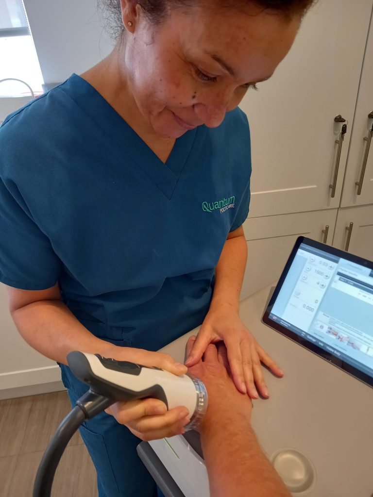 Medical professional in blue scrubs applies a handheld device to a forearm, watching a tablet screen during a procedure in a clinic kitchen.