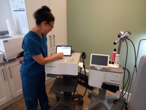 Medical professional in blue scrubs works at a tablet console in a clinic, with medical devices nearby on a cart and counter.