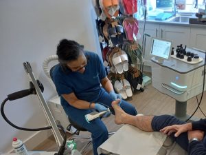 Podiatrist in blue scrubs with gloves examines a patient's foot using a handheld device in a clinical setting with shoe displays in the background.